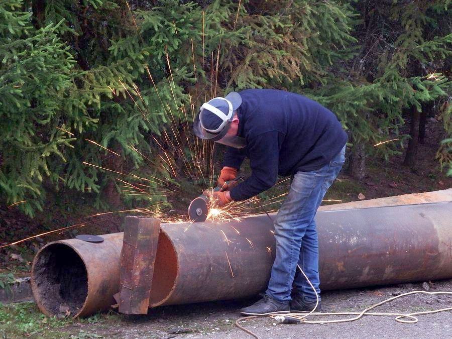 A worker cutting a large steel pipe with an angle grinder